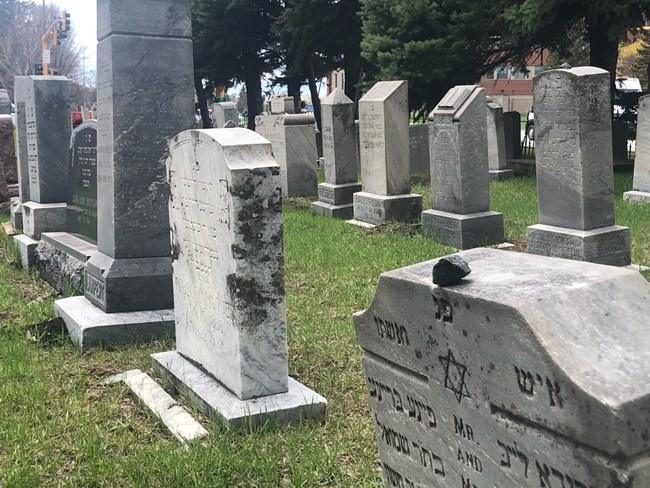 Grave markers arrayed in rows with inscriptions in Hebrew. A small stone has been placed on the marker in the foreground.