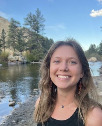 Clara Deforest Colvig Blonde haired blue eyed girl smiling with a river, rocks, and trees in the background.