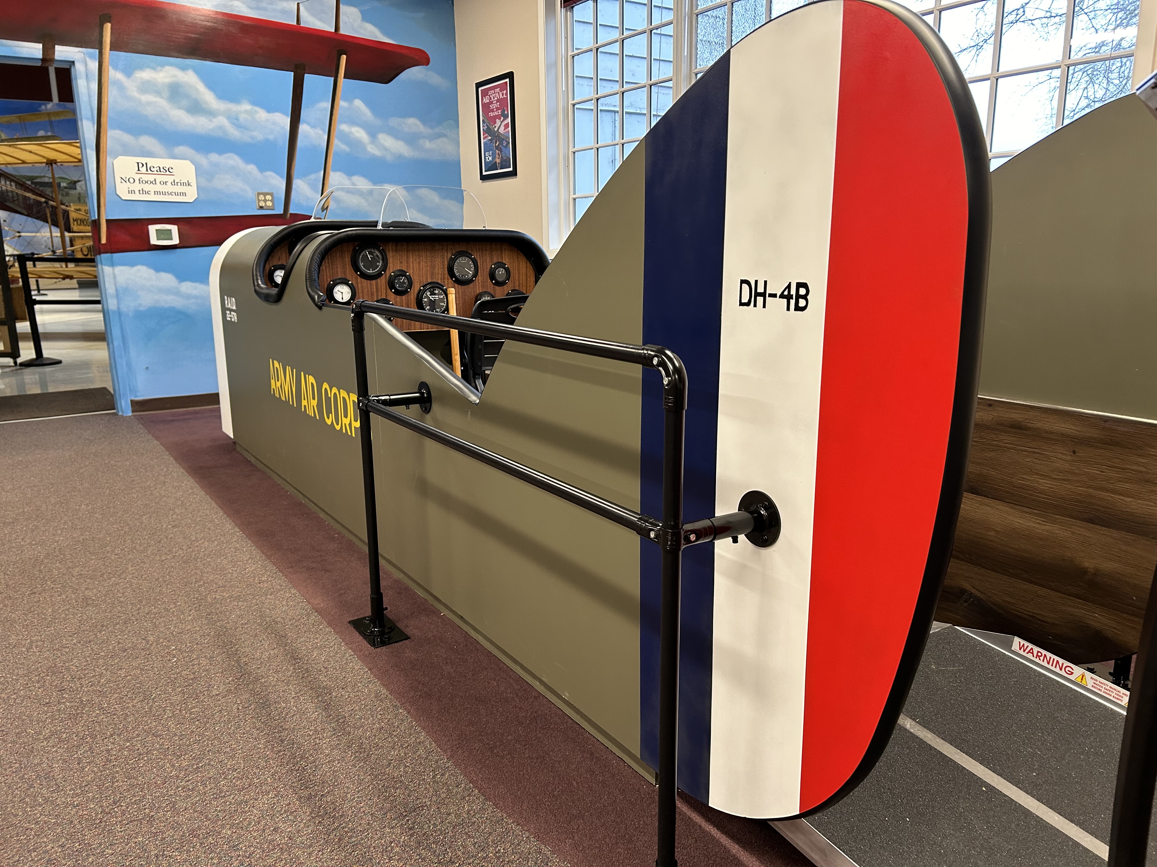 The replica DH-4 Liberty Plane cockpit in the lobby of Pearson Air Museum. It's tail is in the foreground, painted red, white, and blue with "DH-4B" painted on it. The cockpit is olive green with "Army Air Corps" painted on its side in yellow.