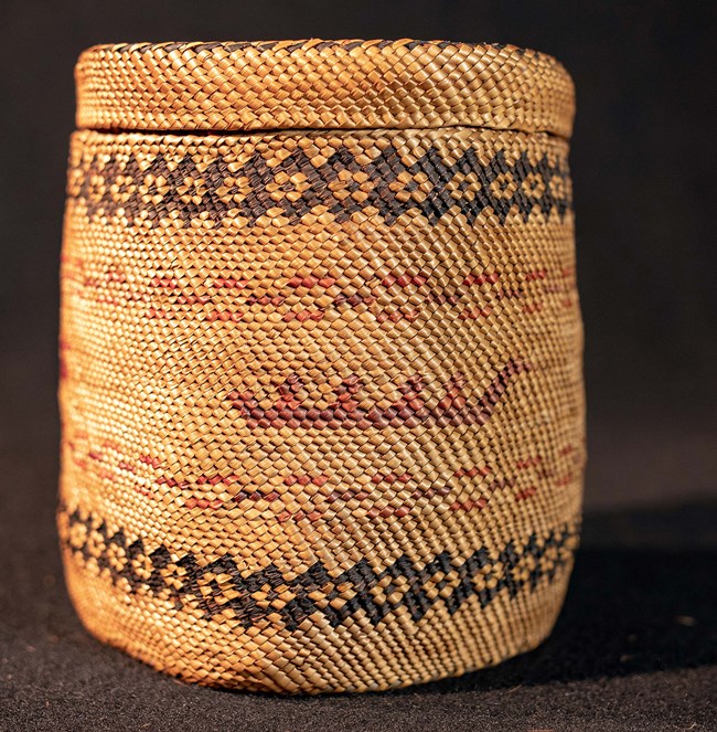 Cylindrical basket woven of natural fibers. Top and bottom bands of black repeating diamonds. In center, design appears to be four people in profile sitting in canoe.