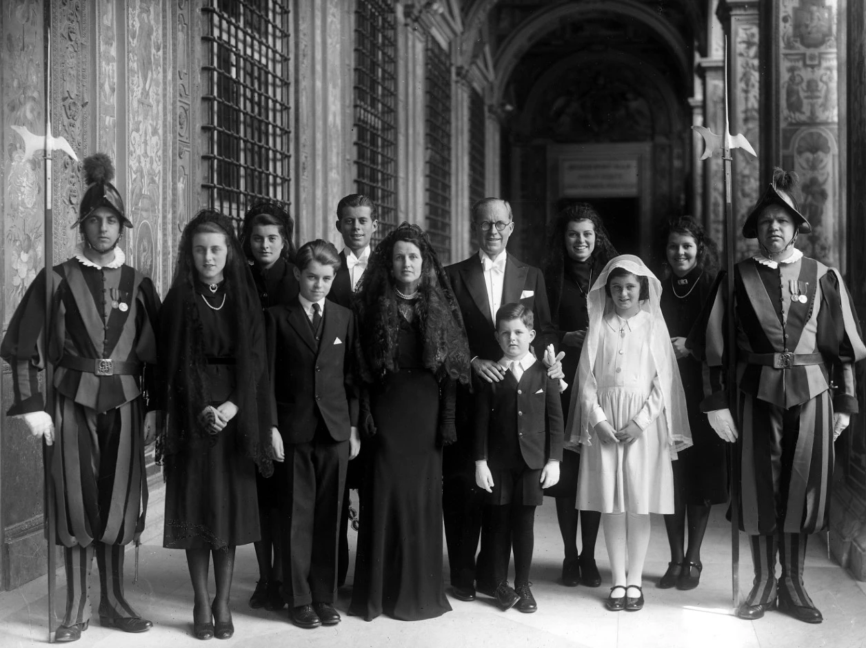 Kennedy family at coronation of Pope Pius XII A black and white photo of a family of ten standing in an arched walkway, between two Swiss Guards in striped uniforms with halberds. The men and boys wear black suits, while the women and girls wear black dresses and veils, except the youngest, in white.