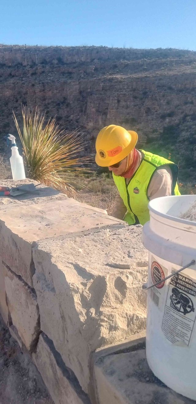 A person is wearing a yellow hard hat, yellow safety vest and dark protective glasses; kneeling behind a tan colored, rectangular stone guardrail next to a yucca plant. A 5 gallon bucket is in the foreground, spray bottle and tools in background.