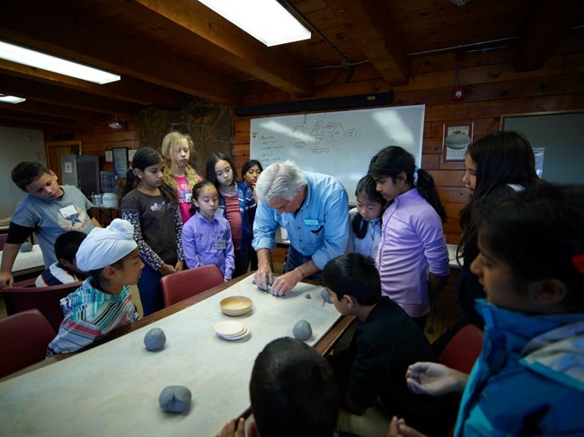 Children surrounding a man showing objects on a table