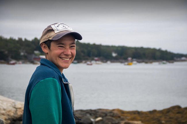 Woman wearing a ball cap and a broad smile stands along the shoreline of a coastal harbor Woman wearing a ball cap and a broad smile stands along the shoreline of a coastal harbor