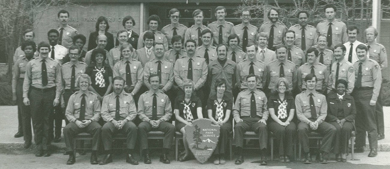 Five rows of employees in different NPS uniforms all facing the camera two women hold a large arrowhead in front of them as they are seated in the front middle. A low building is seen behind them and bare trees frame the group on either side.