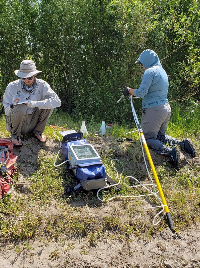 Two scientists sitting on a sandy stream bank collecting water in a sample container from a backpack with a long tube.