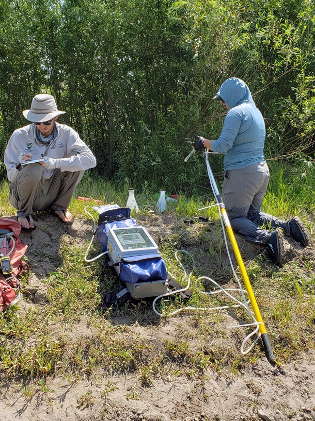 Processing eDNA water samples from a backpack collector. Two scientists sitting on a sandy stream bank collecting water in a sample container from a backpack with a long tube.