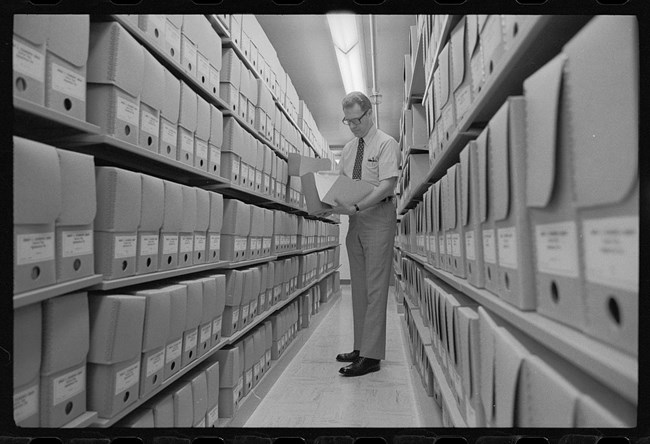 Black and white photo of a man looking at a folder in a row of file boxes