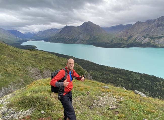 Eric Andre A person stands with two thumbs up on a grassy overlook with a turquoise lake and mountains in the distance