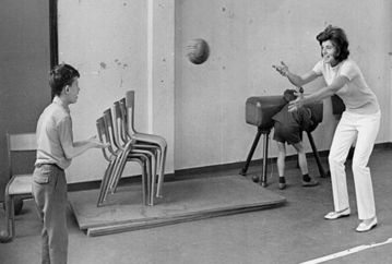 A black and white photo of Eunice Shriver tossing a volleyball to a young boy in a room with chairs and a desk. Another child looks under a desk in the background.