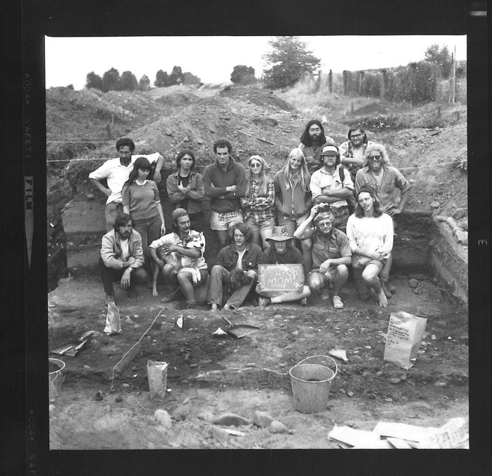 A black and white photo showing a group of men and women wearing 1970s-style clothing near an archaeological excavation area.