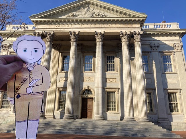 A cartoon illustration cutout of Hamilton is held in front of a marble building with columns.