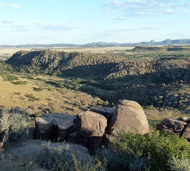 View of rocky desert hills covered in shrubs and valleys with desert plants and buildings.