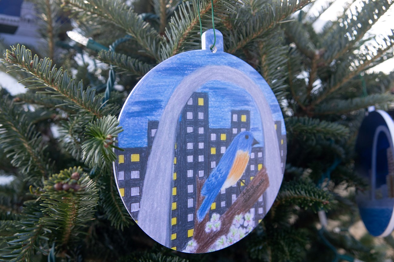 ornament of drawing of an arch with city buildings in the back, and a bird in the middle. The ornament is hanging from christmas tree