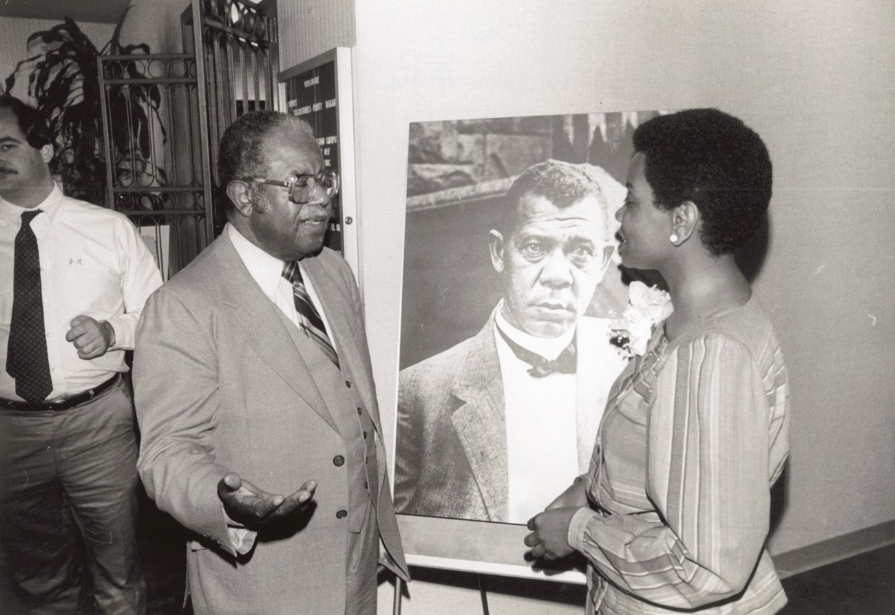 A woman and a man stand chatting in front a photo of Booker T. Washington.