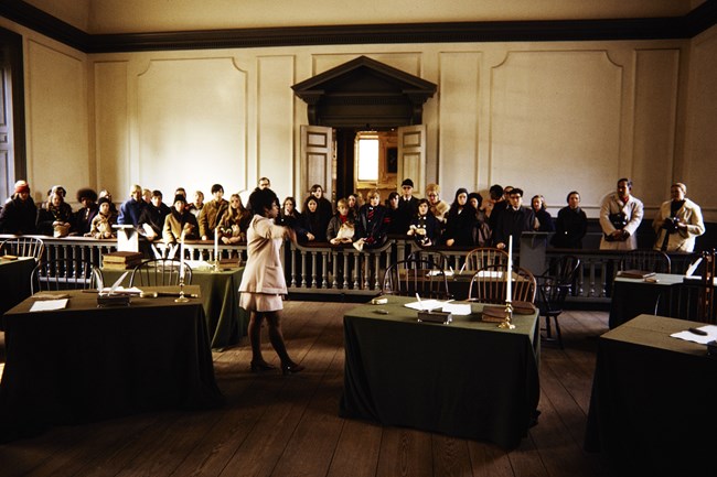 Geraldine Bell in beige NPS uniform stands between two tables addressing visitors standing behind a wooden railing.