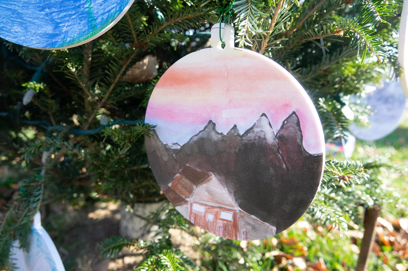 grand teton national christmas tree 2021 ornament with a drawing of mountains with snow and a house, portraying grand teton national park. The ornament is hanging from christrmas tree