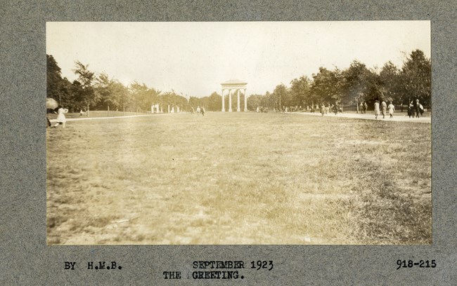 Black and white photograph of large, flat open grassy area with walking paths on both sides, with some people using them. At the end are four large white pillars, with a top, connecting them all