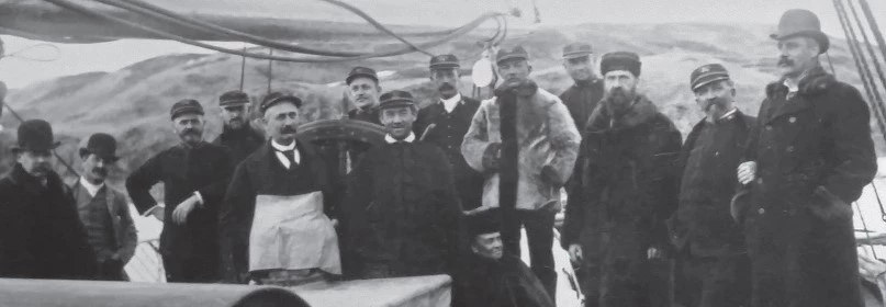 a group of 13 men pose in a historic scene on the deck of a ship. One of them is a Black man wearing a light jacket.