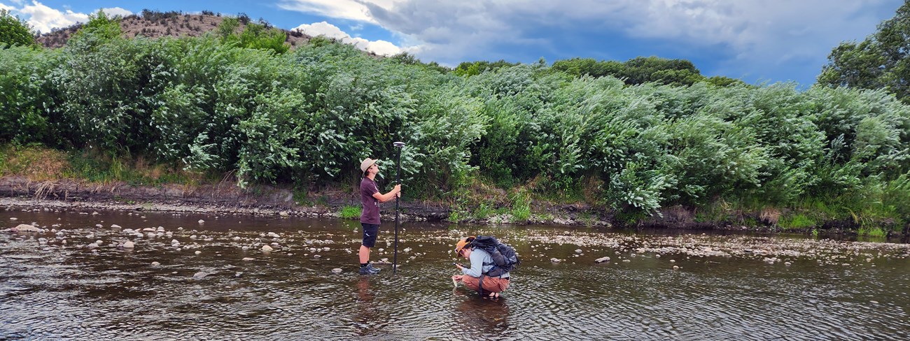 Habitat sampling One scientist bent down to sample water and another scientist holding a water sampling equipment on a long pole attached to a tablet.