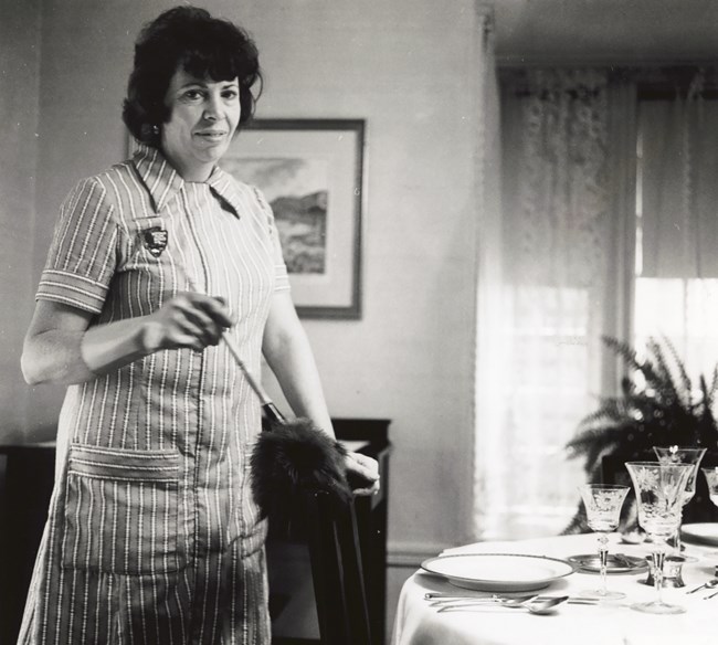Claire Sampson Woman in a smock with a feather duster dusts a set table