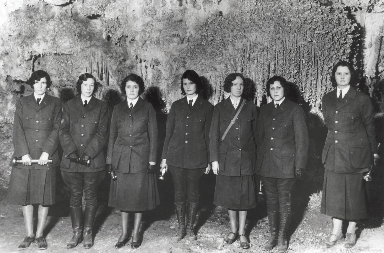 Women at Carlsbad Caverns, 1932 Seven women wearing NPS uniforms stand in a row in front of a rock wall.
