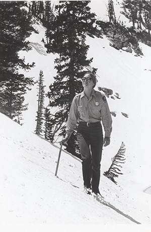 A women wearing a long sleeved shirt and dark pants walks on a mountain towards us with evergreen trees in the background.