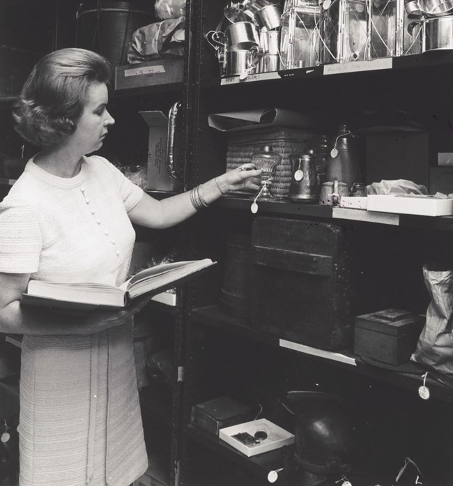 Vera Craig. Vera craig stands in a storeroom holding a ledger in one hand and a lamp in the other.