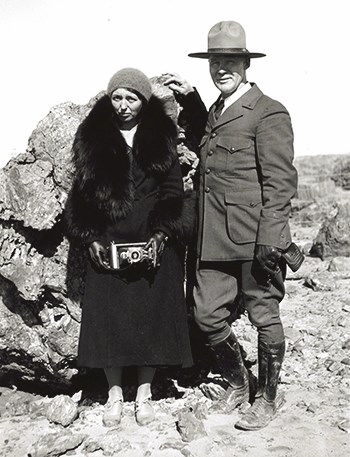 Dama and Charles "White Mountain" Smith. Dama wears a fur coat and holds a camera.
