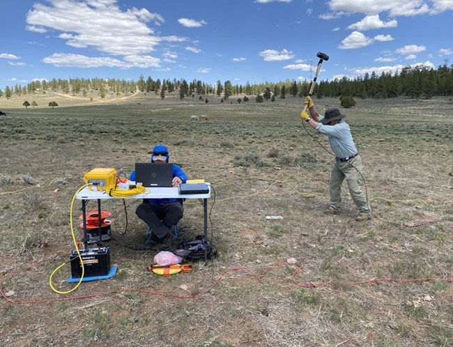 One person swings a hammer, and another person sits behind a computer with some equipment in a field.