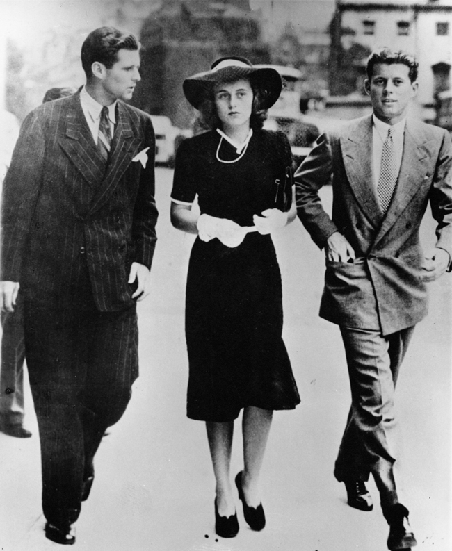 Joe Jr., Kick, and Jack walking to Westminster A black and white photo of a young woman in a hat, dress and gloves walking between two young men in suits on a city street. The man at left speaks, turned toward the woman, who looks ahead nervously. The man at right faces the viewer.