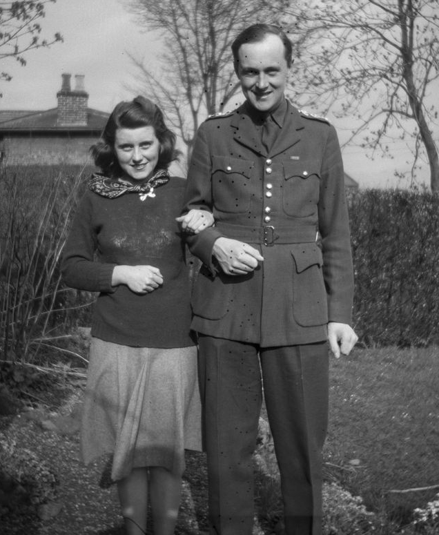 Kick and Billy A black and white photo of a young woman and tall young man standing arm in arm on a lawn. She wears a silk necktie, sweater, and skirt, and he wears a khaki service dress military uniform. Behind them are hedges, trees, and a brick cottage.