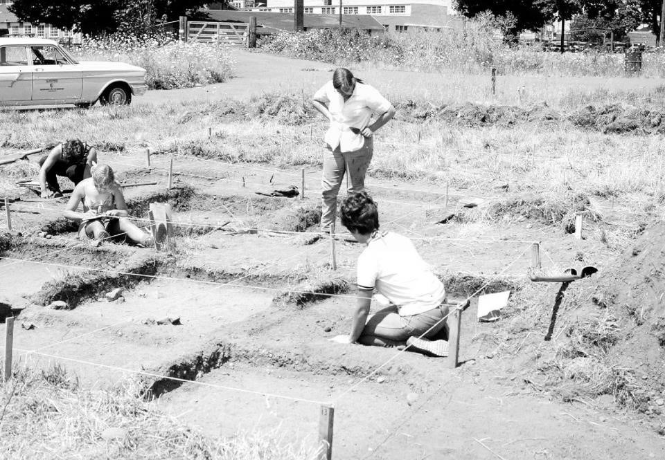 Black and white photo of a woman standing overseeing other women archaeologists working on an archaeological excavation.