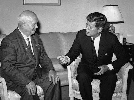 Kennedy and Khrushchev in Vienna The two men are seated in chairs. Khrushchev is on the left and Kennedy is on the right. A couch and lamp are behind them.