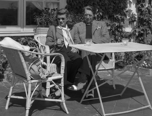 Kick and Peter A black and white photo of a smiling young woman in a blazer, neck scarf, slacks, black flats and sunglasses sitting with a smiling man in a blazer, slacks and black toed white shoes at a white patio table, in front of a building covered in ivy leaves.