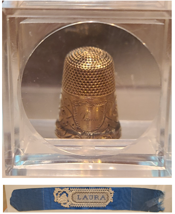 One gold thimble with the name “Laura” etched on a shield and surrounding designs. Blue bookmark held on white background paper with the name “Laura” stitched onto a white doilie-esque center.