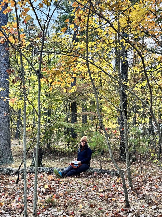 A woman sits in a colorful forest, sketching in a sketchbook.