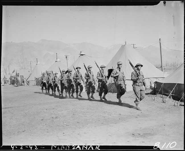 Soldiers with guns march in a single file line in front of army tents