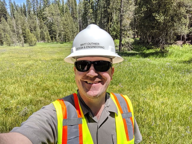 Selfie of a man wearing a hardhat, which says Matt Outhier Design & Engineering, and a safety vest over a ranger uniform with a meadow and forest in the background