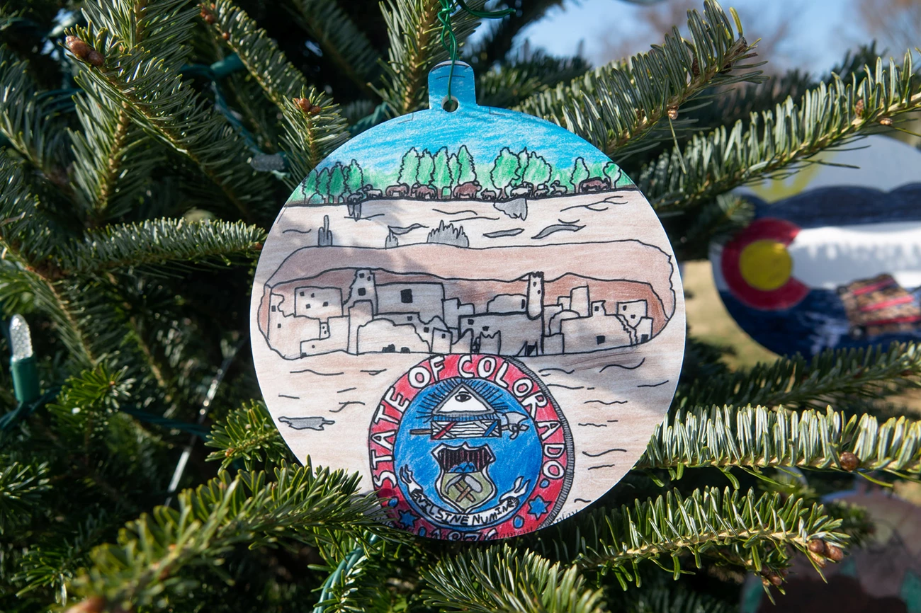 mesa verde national christmas tree 2021 ornament of a drawing of mesa verde's archeological site. The ornament is hanging from christmas tree