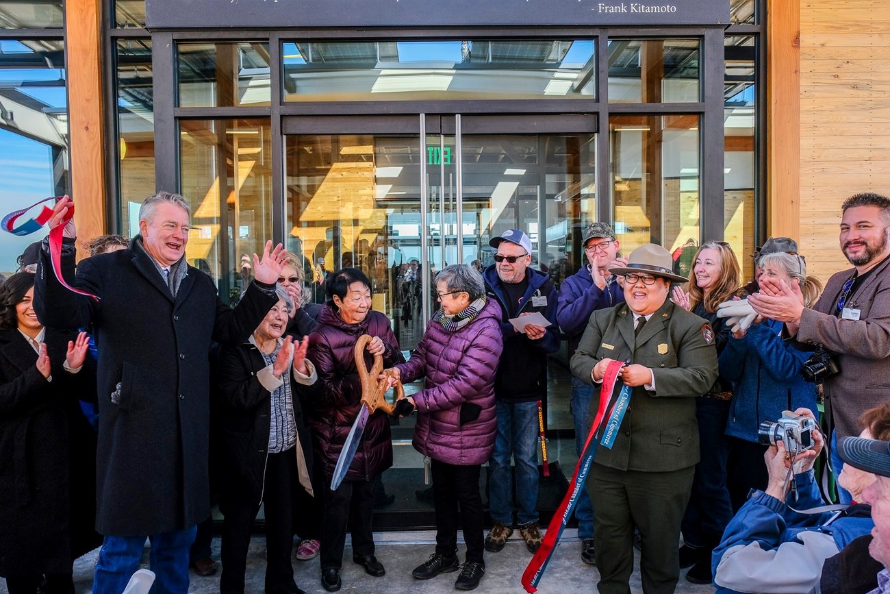 A group of smiling people, including a park ranger, cut the ceremonial ribbon outside the new Minidoka visitor center.