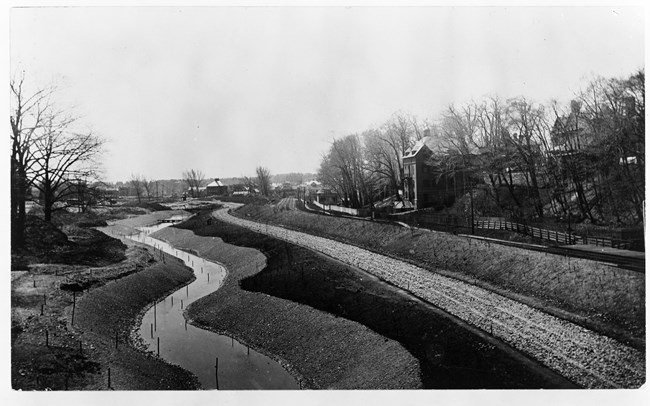 Black and white photograph of river being dug out of ground, with hills going up on both sides. Both sides have trees but no leaves, and on one side is a line of homes.