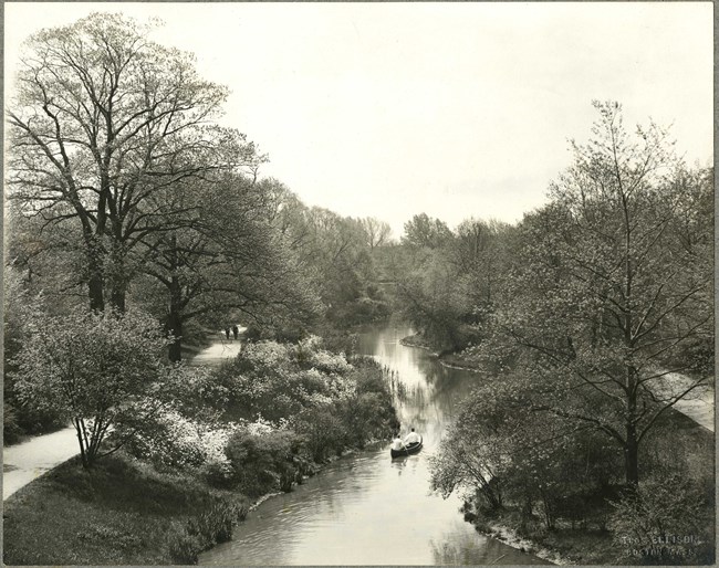 Black and white photograph of body of water with two walking paths and shrubs on both sides. There is a canoe in the middle of the water.