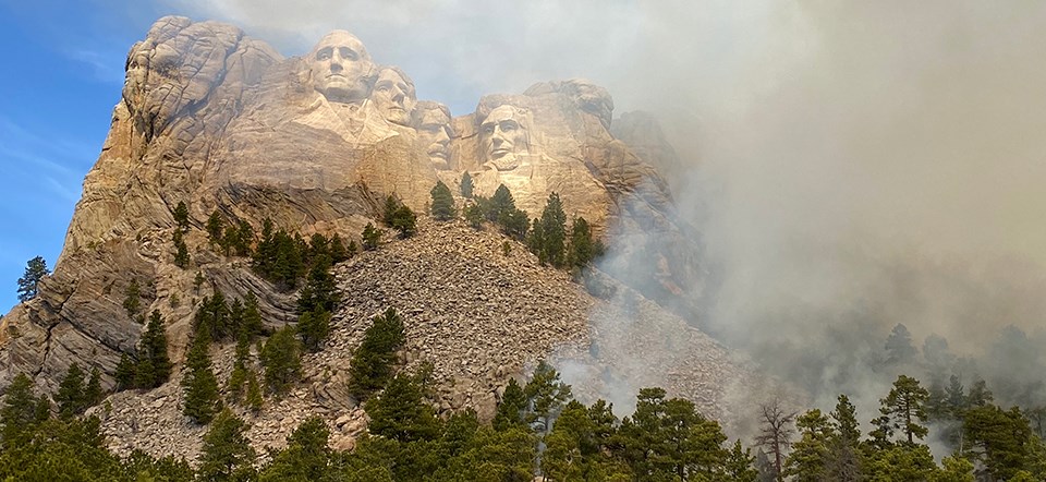 Smoke rises from trees on slope below carved figures on Mount Rushmore, obscuring view. Smoke rises from trees on slope below carved figures on Mount Rushmore, obscuring view.