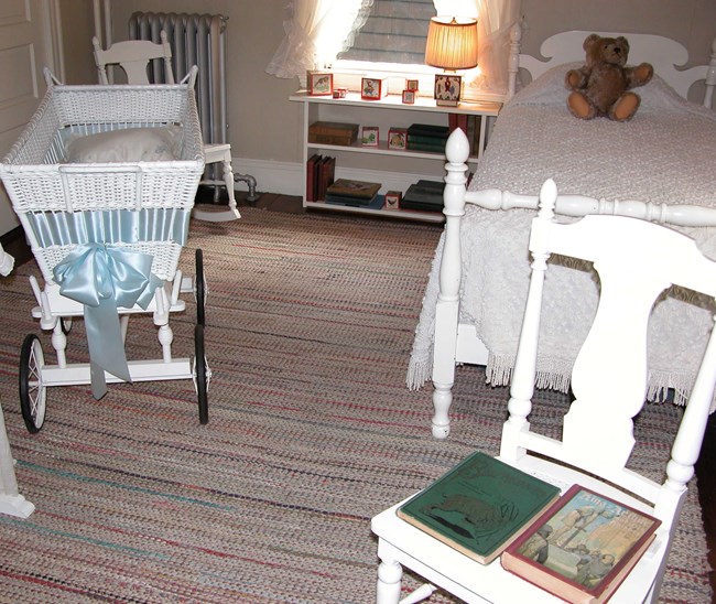A photo of a nursery with white furniture: a crib, a shelf with a lamp and toy blocks, a bed with a teddy bear, and a chair with two books on it. One’s cover is green with a jumping goat, the other is red with a knight pulling a sword from a stone.