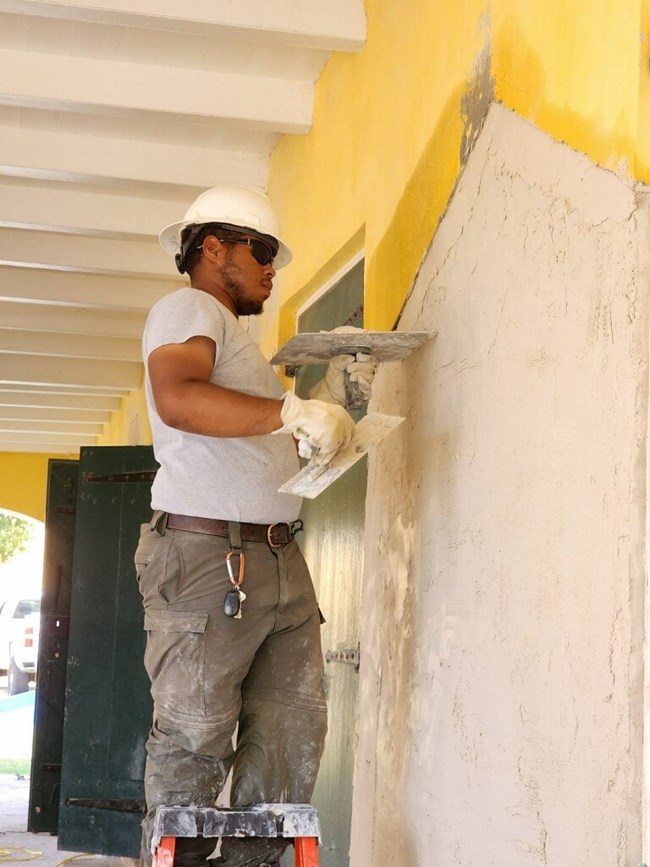 Man standing on latter with a hard hat applies plaster to exterior building.