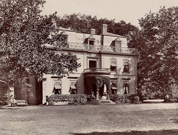 Black and white photograph of large brick home with many terraces and vines creeping over it.