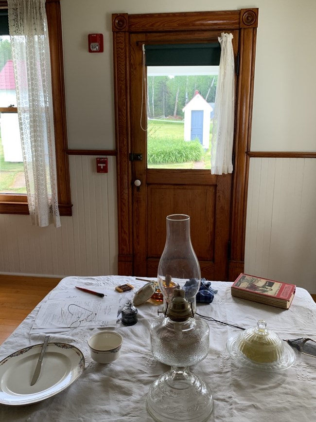 A kitchen table set with a white table cloth, dishes, lantern, and book looking through a glass window at a small outside outhouse.