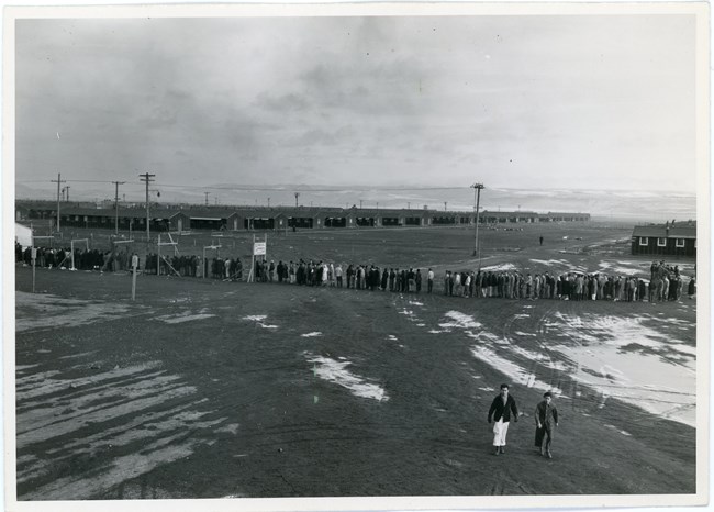 Long lines of people wait across an empty dirt area with wooden buildings in the background