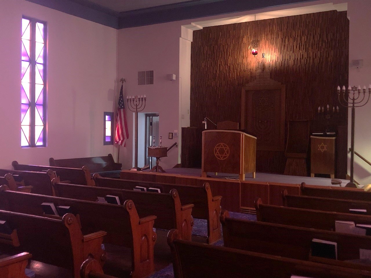 Interior of a synagogue illuminated by a stained glass window. The bimah includes a centered reading table, a suspended lamp, and a textured wall.